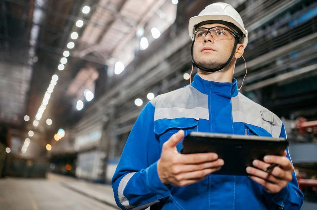 Operaio con casco e tablet in una fabbrica illuminata, concentrato sul lavoro.